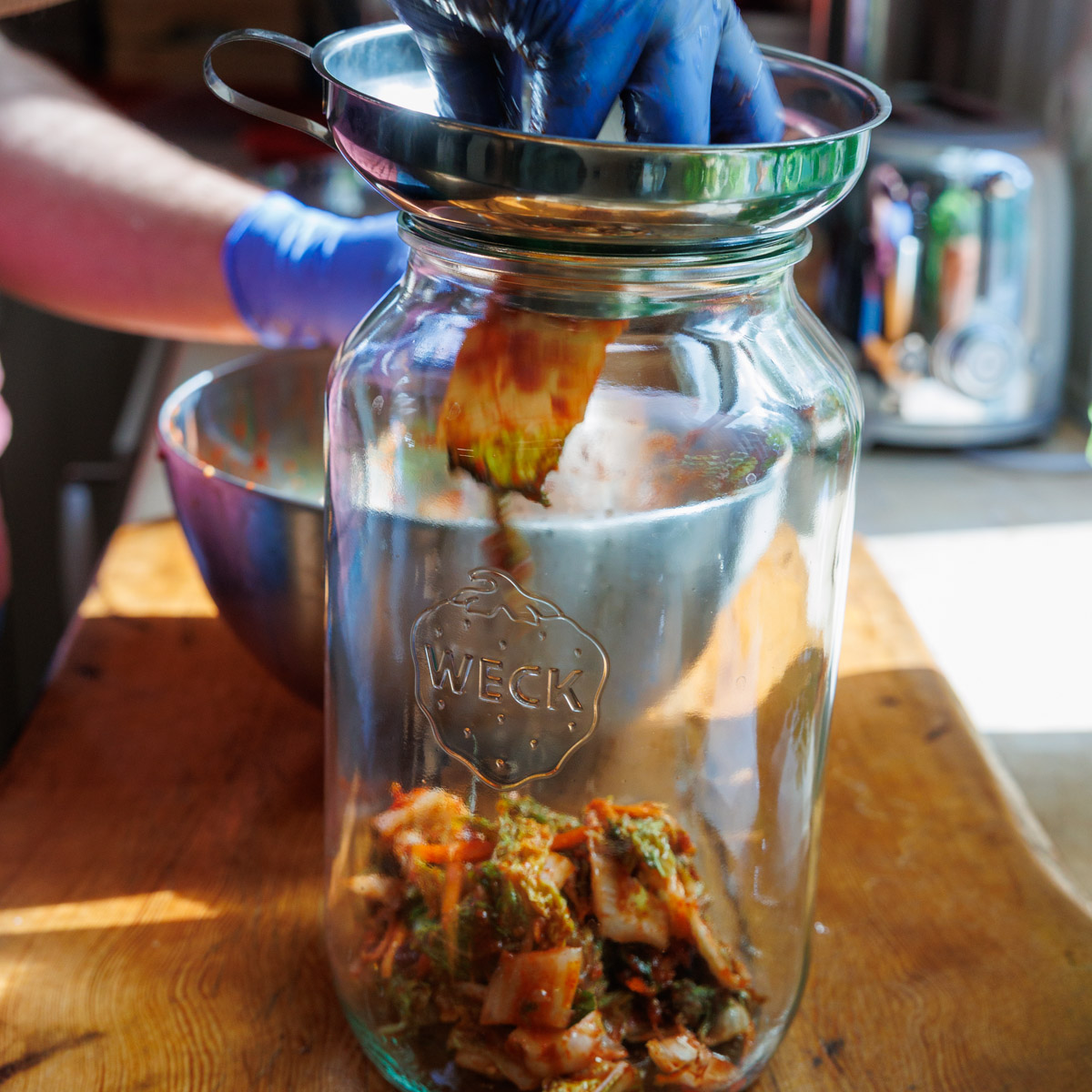 Kimchi fylls i glasburk med tratt och handskar
